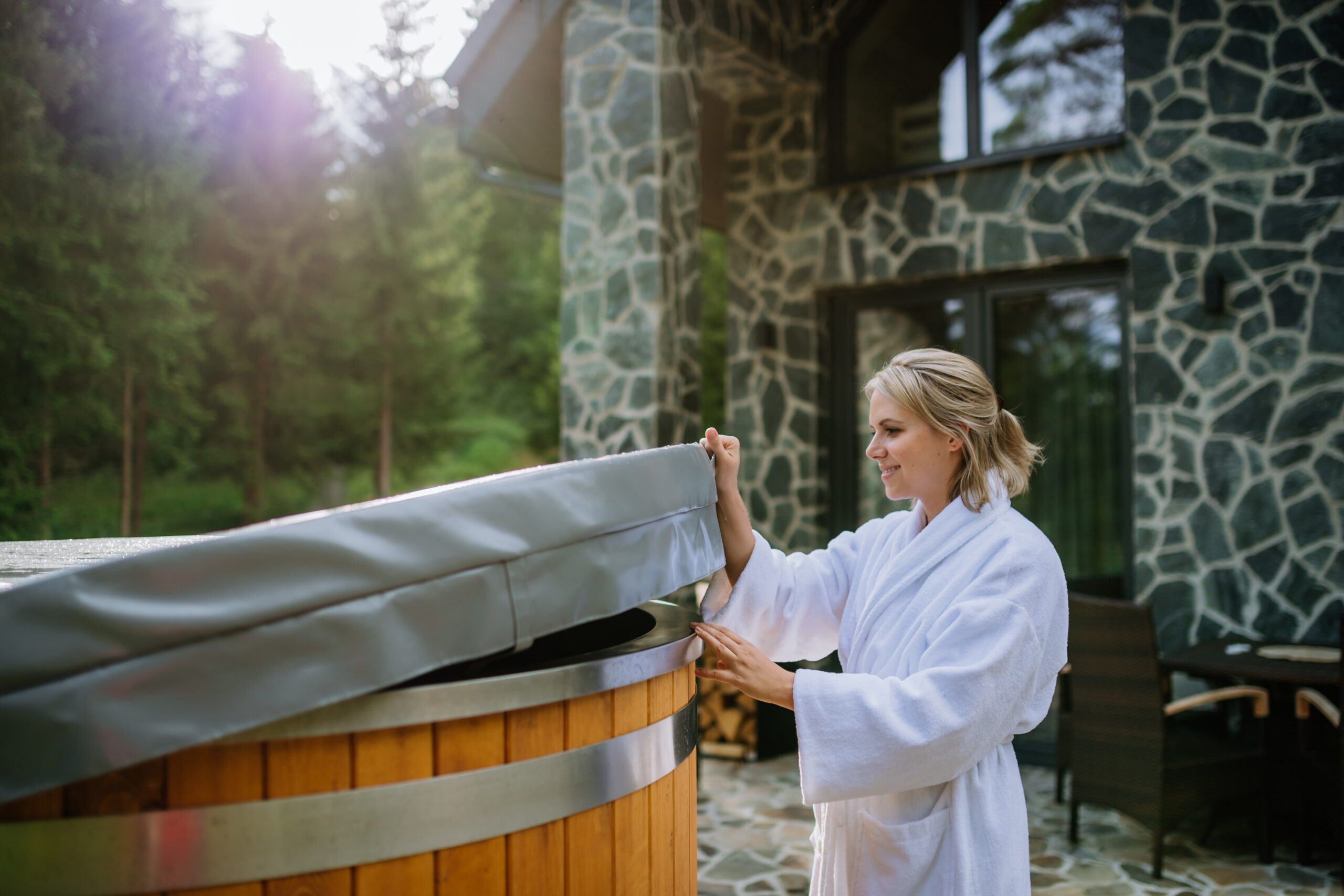 A woman in bathrobe opening lid of hot tub, checking temperature, ready for home spa procedure in hot tub outdoors. Wellness, body care, hygiene concept.